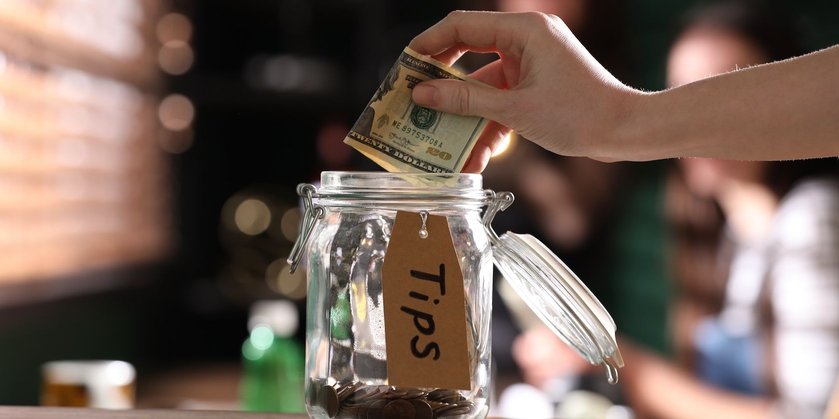 A hand placing several dollar bills into a glass jar labelled “tips”