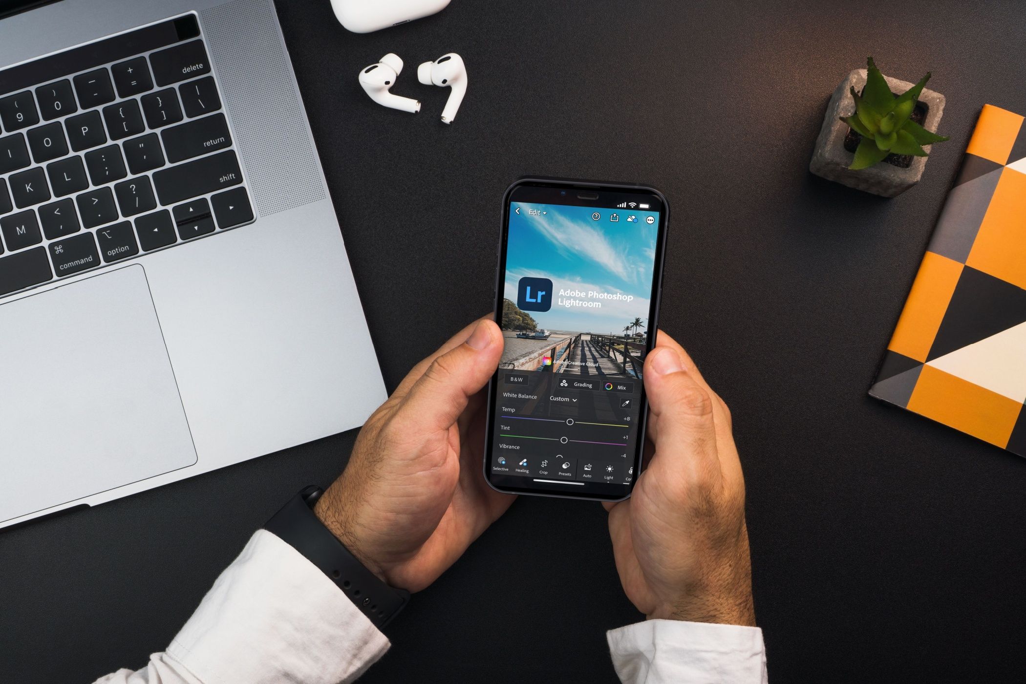 Man holding smartphone displane Adobe photo editing app, with laptop, earbuds, and notebook in background.