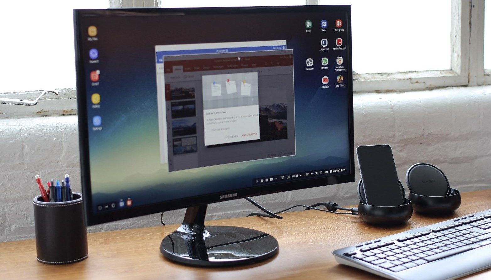 The Samsung monitor is on a desk with a keyboard and sound system