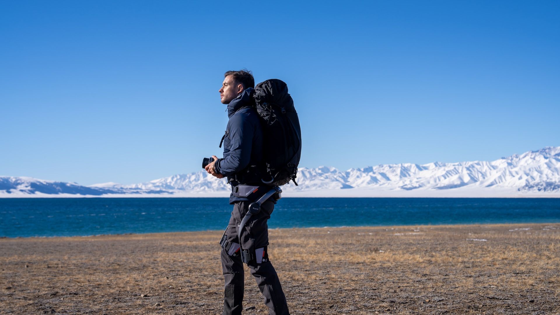 A man holding a camera wearing a Hypershell X with snowy hills and a lake in the background.