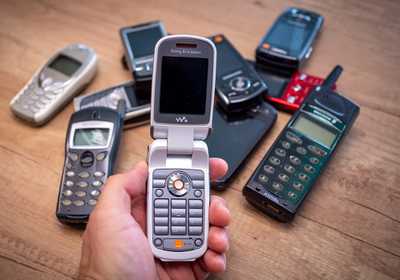 Person holding an old Sony Ericsson flip phone in front of a group of various vintage mobile phones