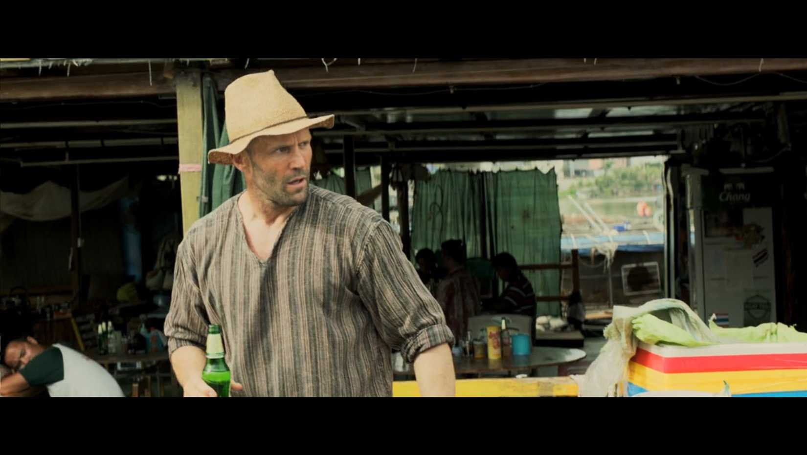 Jason Statham standing, wearing a hat, and holding a beer