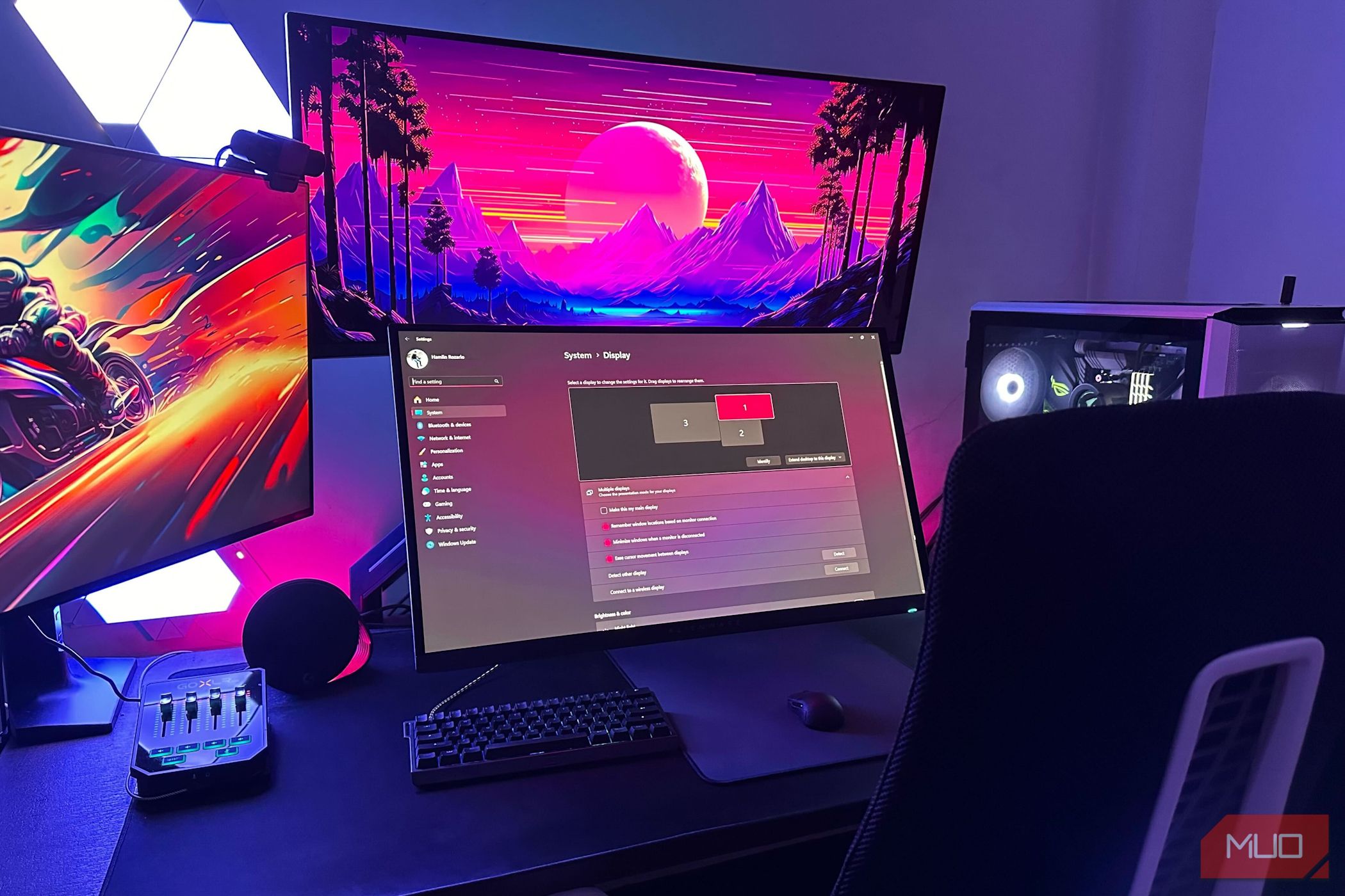 Three gaming monitors mounted on a computer desk with keyboard, mouse, and a chair