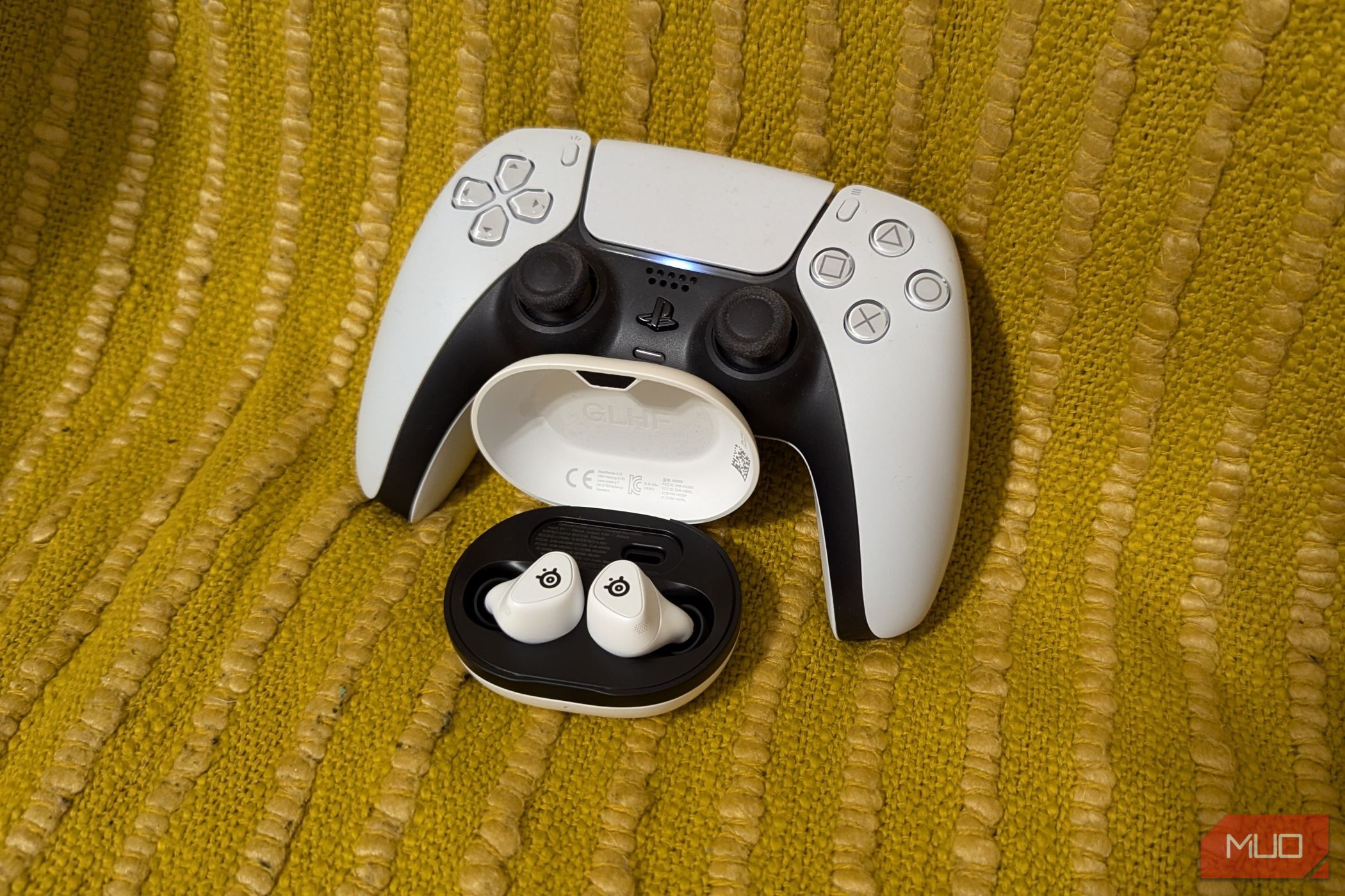 a PlayStation 5 controller with the Arctis GameBuds in front, resting on a yellow cloth